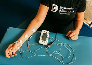 A therapist's arm with electrodes attached and a stimulation unit on a teal table