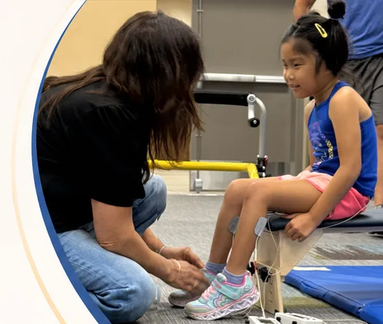A therapist adjusting electrode leads on a child's leg while the child sits on a therapy bench